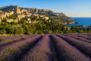 Panoramic view of a lavender field leading to a perched stone village with Alpilles mountains and a distant calanque under a golden Mediterranean sky.